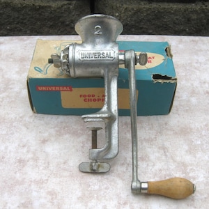 May include: A silver-colored metal Universal food chopper with the number 2 embossed on the top. It features a hand crank with a light brown wooden handle and a screw-style clamp base. The chopper is positioned in front of its original blue box.