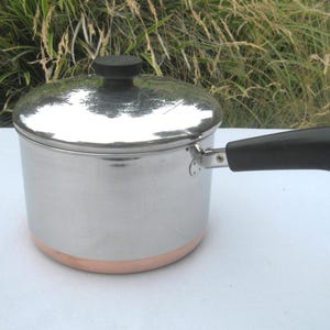 May include: A stainless steel saucepan with a copper-colored base, a matching lid with a black knob, and a black handle featuring a small metal hanging loop. The pot has a polished, reflective finish and a cylindrical shape with a flat bottom.