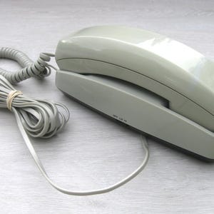 May include: A vintage, light gray telephone with a coiled handset cord and a straight cord. The phone has a rectangular base and a curved top. The cords are bundled with a rubber band. The phone is a classic landline design.