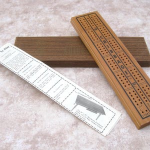 Könnte beinhalten: Ein hölzernes Cribbage-Spielbrett mit dem Aufdruck "CRIBBAGE", eine Holzbox und ein Papier mit Anweisungen. Das Brett hat Löcher für die Stifte und ist aus hellbraunem Holz gefertigt.