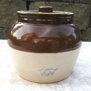 May include: A two-toned ceramic crock with a dark brown glazed upper section and a cream-colored lower section. The vessel features a matching lid with a small knob and a blue stamped crown logo containing the number 3 on the front lower body.