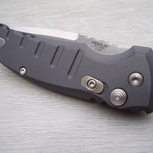 May include: A gray folding knife with a partially serrated blade. The knife has a textured handle with recessed grooves and silver-colored hardware. The blade is marked with "Hogue A01-MS USA".