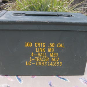 May include: An olive drab metal ammunition box with a hinged lid and a black handle. The box has yellow stenciled text that reads "100 CRTG .50 CAL LINK M9 4-BALL M33 1-TRACER M17 LC-09B614L653".