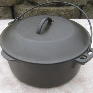 May include: A black cast iron Dutch oven with a lid and a handle. The pot has a rounded shape with a handle on the lid and a wire handle for carrying. The Dutch oven is designed for cooking.