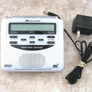 May include: A white Midland weather alert radio with a black digital display, control buttons, and a speaker grille. The device features a retractable antenna and includes a black power adapter with a cord. Text includes WARNING, WATCH, ADVISORY, MENU, SELECT, UP, DOWN, VOLUME, WEATHER/SNOOZE, and S.A.M.E. DIGITAL WEATHER/ALL HAZARDS ALERT RADIO.