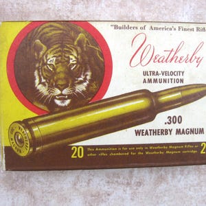 May include: A rectangular box of Weatherby .300 Weatherby Magnum ultra-velocity ammunition. The yellow and white packaging features a tiger illustration in a red circle, the text Builders of America's Finest Rifles, and the number 20.