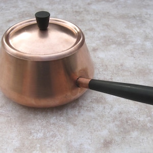May include: A copper pot with a matching lid and a long, straight black handle. The lid features a small, cylindrical black knob at the center. The pot has a rounded, tapered body and a smooth, metallic finish throughout the exterior surface.