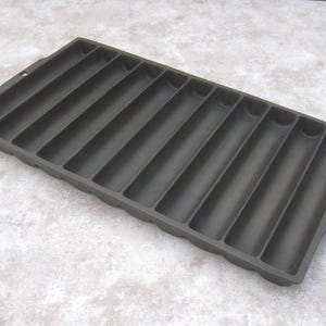 May include: A dark gray cast iron cornbread pan with 12 individual molds. The rectangular pan has a handle on one end and a small hole for hanging. The interior of the pan has a non-stick surface.