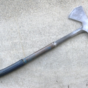 May include: A metal hatchet featuring a silver-toned steel head with a curved blade and a flat poll. The handle is constructed from a long, dark, weathered metal shaft with a black textured grip at the base, showing signs of use.