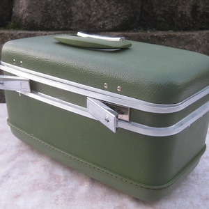 May include: A rectangular olive green train case with a textured exterior finish. The case features a top handle with silver-tone accents, a silver-tone metal frame, and two side latches that are currently in the open, extended position.