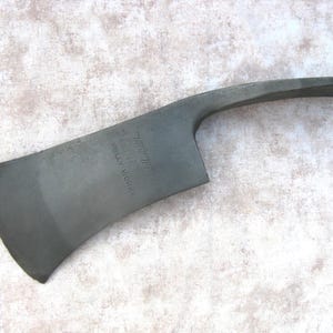 May include: A dark grey metal fire axe head featuring a curved blade and a pointed pick end. The surface of the metal is stamped with the text True Temper Kelly Works. The tool head has a smooth, matte finish and a distinct, functional shape.