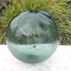 May include: A large, round, clear glass float with a slight green tint. The float has a smooth surface and is partially filled with water. The background features green and yellow grass and a stone wall.