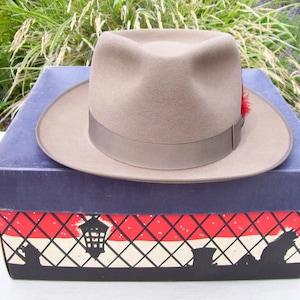 May include: A tan fedora hat with a brown band and a red feather accent, resting on a blue box with a decorative red and white pattern. The hat has a classic, vintage style.