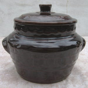 May include: A dark brown ceramic crock pot with a matching lid. The pot has two handles and a textured surface. The lid has a small knob and a decorative wavy pattern around the edge. A classic kitchenware item.