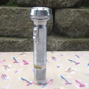 May include: A vintage silver flashlight with a ribbed body and a chrome head. The flashlight has a switch on the side and a small label near the bottom. The background is a stone wall and a table with a floral pattern.