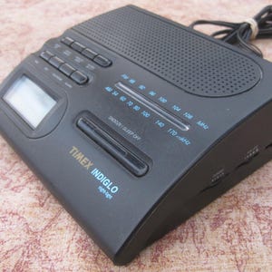 May include: Black Timex Indiglo clock radio with a digital display and AM/FM radio tuner. The radio has a sleep timer and snooze button. The clock radio has a black and silver design.