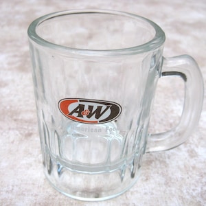 May include: Clear glass A&W mug with a handle. The mug features the A&W logo in red, black, and white. The mug has a rectangular shape with vertical indentations and a sturdy handle.