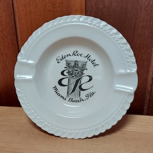 May include: White ceramic ashtray with a black and white design of the Eden Roc Hotel in Miami Beach, Florida. The design features a column with the hotel's initials.