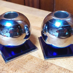 May include: A pair of spherical candle holders featuring a polished chrome finish. Each sphere is mounted on a square, deep blue glass base with a thin silver-toned metal border, creating a reflective surface that mirrors the surrounding environment.