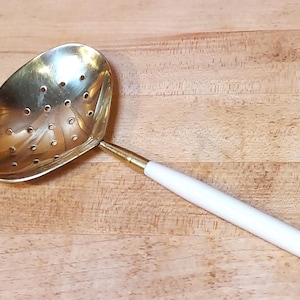 May include: A brass-colored slotted spoon with a white handle. The spoon has a rounded, oval bowl with multiple small holes. The handle is white with a brass-colored band where it connects to the spoon.