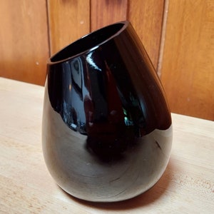 May include: A black, glossy, teardrop-shaped vase with a wide opening. The vase is sitting on a wooden surface.