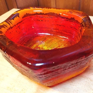 May include: A square, glass bowl with a textured exterior. The bowl features a gradient of colors, transitioning from a deep red at the base to a vibrant orange and yellow at the top. The rim has a darker, textured appearance.
