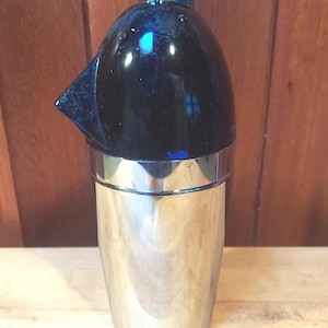 May include: A vintage cocktail shaker with a black top and a polished silver base. The top features a built-in spout and a small cap. The shaker is designed for mixing drinks and has a classic, retro aesthetic.