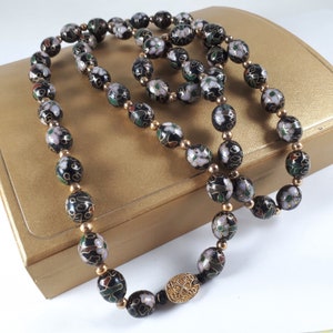 May include: A long black and white beaded necklace with gold accents. The beads are oval shaped and have a floral design. The necklace is on a gold box.
