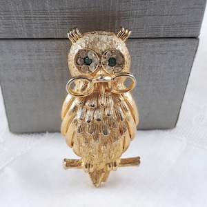 May include: A gold-tone owl brooch with green and clear rhinestones for eyes. The owl is perched on a branch and has a bow tie.