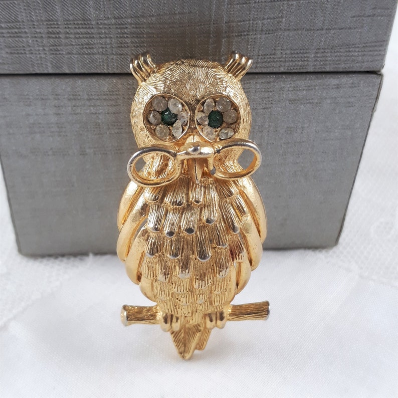 May include: A gold-tone owl brooch with green and clear rhinestones for eyes. The owl is perched on a branch and has a bow tie.