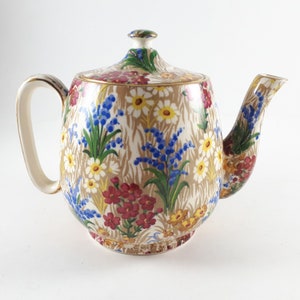 May include: A white teapot with a floral pattern in shades of blue, red, yellow, and green. The teapot has a rounded body and a spout on the side.