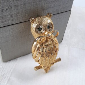 May include: A gold-tone owl brooch with green and clear rhinestone eyes. The owl is perched on a branch and has a bow tie.
