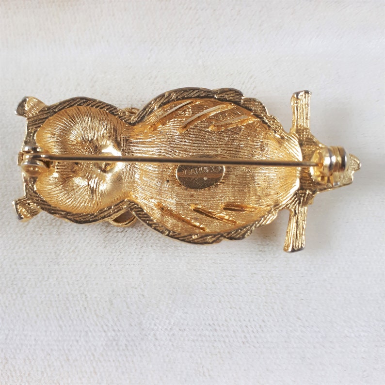 May include: Gold-toned owl brooch with a textured design and a pin on the back. The brooch is marked "TANGELO" on the back.