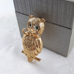 May include: A gold-tone owl brooch with green and clear rhinestones for eyes. The owl is holding a pair of glasses in its beak.