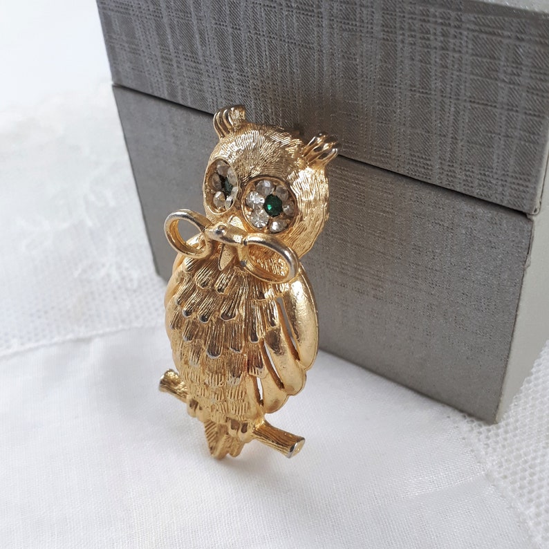 May include: A gold-tone owl brooch with green and clear rhinestones for eyes. The owl is holding a pair of glasses in its beak.