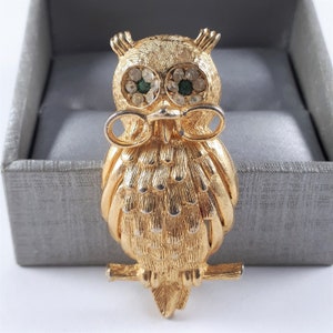 May include: A gold-tone owl brooch with green rhinestone eyes. The owl is perched on a branch and has a detailed feathered design.