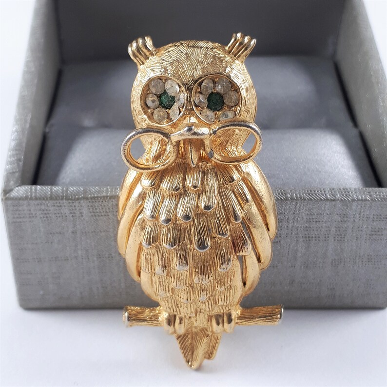 May include: A gold-tone owl brooch with green rhinestone eyes. The owl is perched on a branch and has a detailed feathered design.