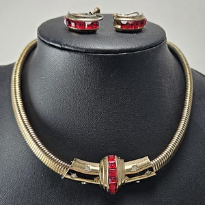 May include: A vintage gold-tone necklace and matching earrings set. The necklace features a flexible, ribbed design with a central rectangular red stone cluster. The earrings are small hoops with matching red stones. The jewelry is displayed on a black mannequin.