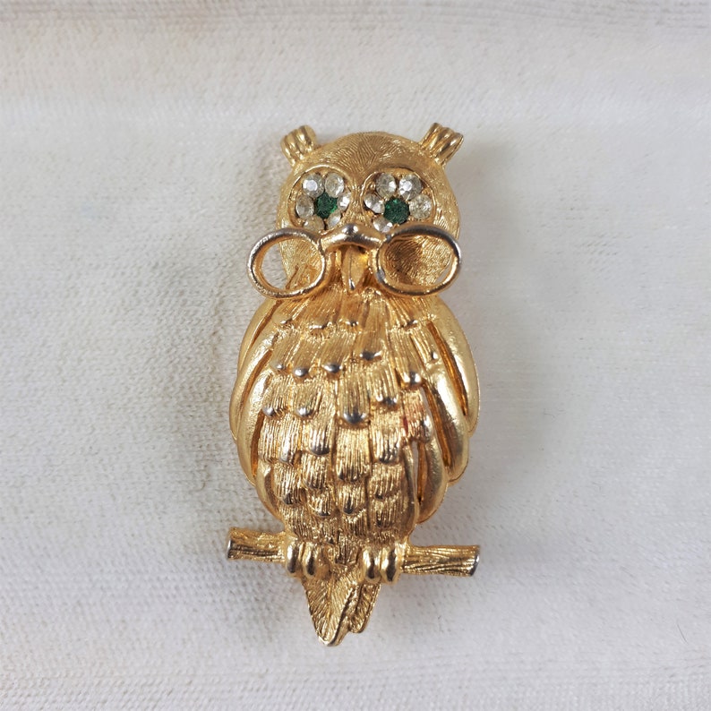May include: A gold-tone owl brooch with green and clear rhinestones for eyes. The owl is wearing glasses and perched on a branch.