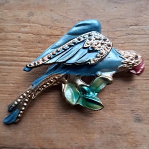 May include: A blue and gold bird brooch with a red beak perched on a green leaf. The bird has a detailed wing and tail with gold accents.