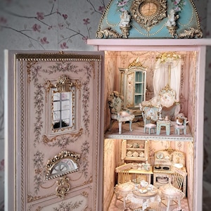 May include: A miniature dollhouse cabinet with a light pink exterior, gold ornate trim, and a blue domed top. The interior features two levels with detailed furniture, including chairs, tables, a bed, and kitchen items. The text AIDA PRAVIA is visible.