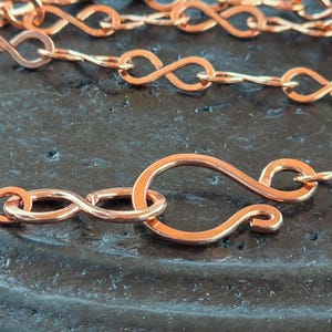 Puede incluir: Primer plano de un collar de color cobre con un diseño de cadena de símbolo de infinito. El collar presenta un cierre de gancho. La cadena y el cierre están hechos de un metal brillante de color marrón rojizo.