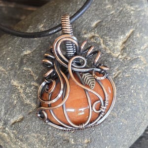 May include: A handcrafted pendant featuring an orange gemstone encased in copper wire, shaped like a pumpkin. The pendant includes a small leaf accent and hangs from a black leather cord. The wirework has a detailed, artistic design.