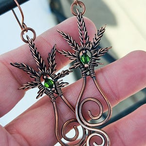 May include: Copper wire earrings with a green crystal in the center of each earring. The earrings are shaped like a marijuana leaf with a dangling spiral.