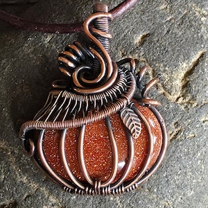 May include: A handcrafted pendant featuring a pumpkin design. The pumpkin is made of a sparkling orange-brown stone, encased in copper wire. The wire is intricately woven, with a leaf detail and a spiral at the top. The pendant hangs from a brown leather cord.
