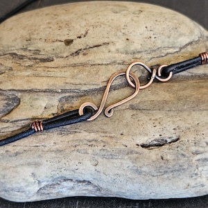 May include: Close-up of a copper clasp on a black leather cord. The clasp is a simple, handmade design with a loop and a hook.