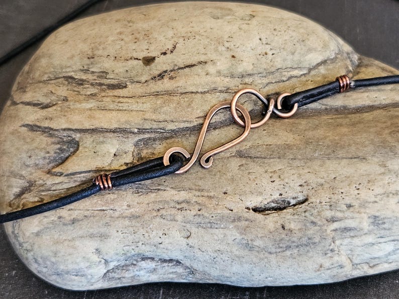 May include: Close-up of a copper clasp on a black leather cord. The clasp is a simple, handmade design with a loop and a hook.
