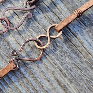 May include: A close-up of a brown leather cord with a copper wire wrap. The cord is attached to a copper wire loop with a decorative infinity symbol design.