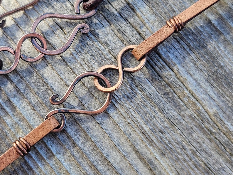 May include: A close-up of a brown leather cord with a copper wire wrap. The cord is attached to a copper wire loop with a decorative infinity symbol design.