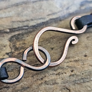 May include: A close-up of a copper-coloured metal clasp with an infinity symbol design. The clasp is attached to a black leather strap.